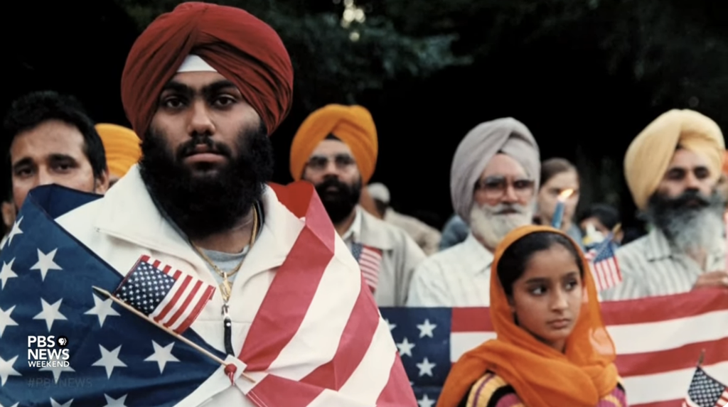 Group of Sikh Americans, including a man wrapped in an American flag and a girl in traditional attire, holding small U.S. flags during a patriotic gathering. The image highlights the Sikh community's pride and integration into American society.