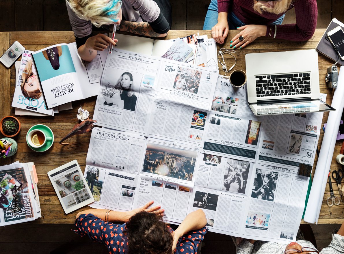  A group of people gathered around a wooden table, analyzing newspapers, magazines, and digital content on a laptop and tablet. The workspace is filled with coffee, notebooks, and creative tools, symbolizing media literacy, news analysis, and digital research.