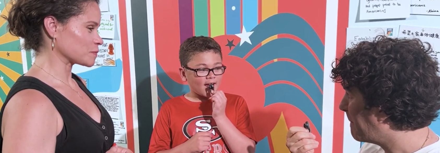 A child stands between two adults, thoughtfully holding a pen near his mouth, as they discuss ideas in front of a colorful wall filled with written hopes and wishes for the future.