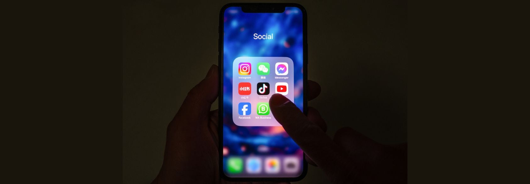 A person holds a smartphone displaying a folder labeled "Social" with popular social media app icons, including Instagram, TikTok, Facebook, YouTube, and Messenger, against a blurred, colorful background.