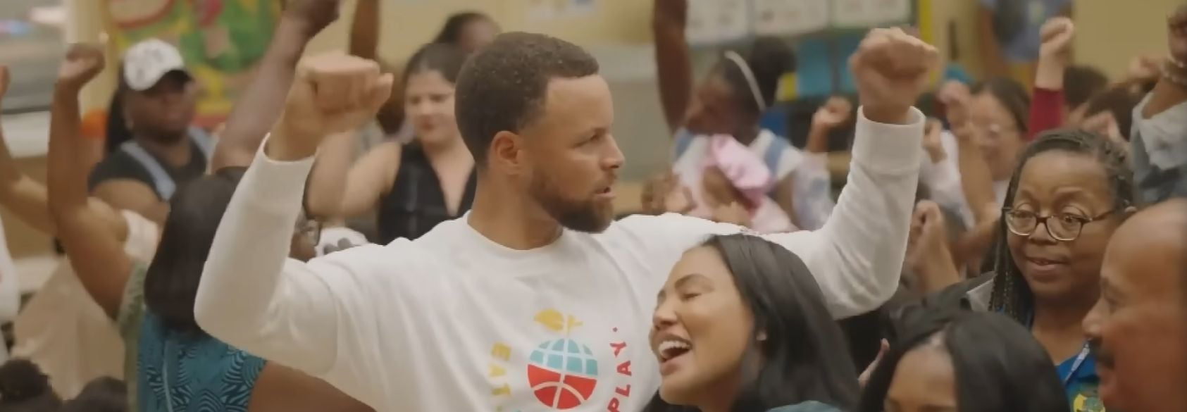 Basketball player Stephen Curry raises his arms in celebration while surrounded by community members at a service event, highlighting efforts with Martin Luther King III to unite communities through service.