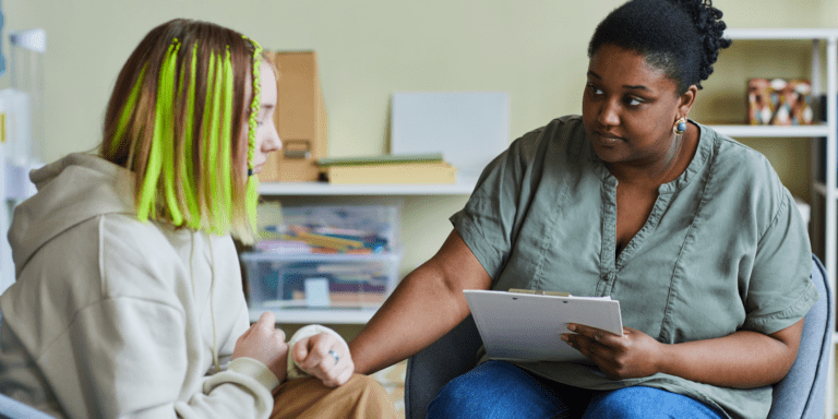 A school counselor sits with a concerned expression while holding a clipboard and offering support to a distressed student with neon green hair, who appears upset. They are seated in a calm, classroom-like setting, suggesting a private and supportive conversation.        Ask ChatGPT